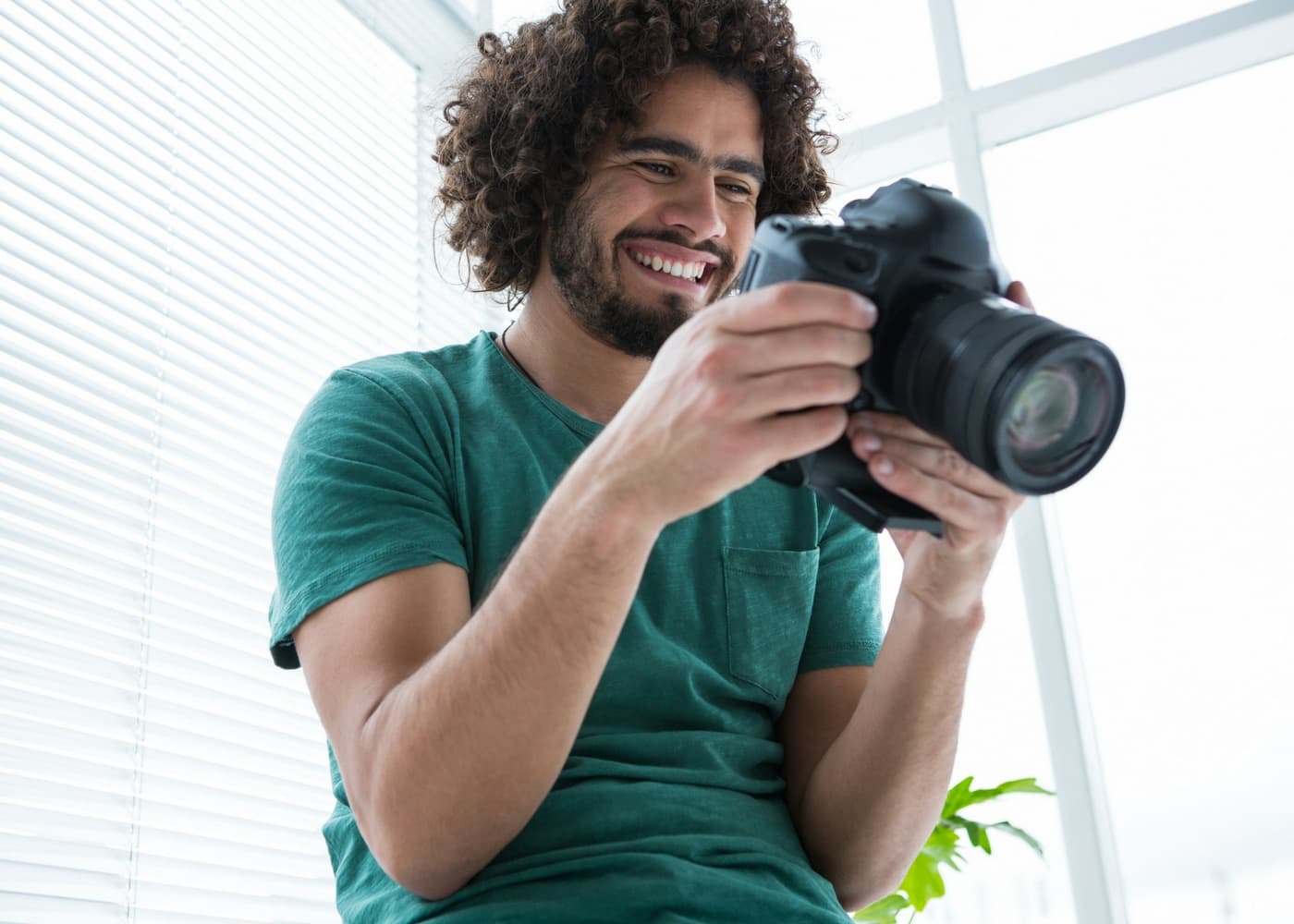 A person smiling at the screen of a DSLR camera beside a bright window