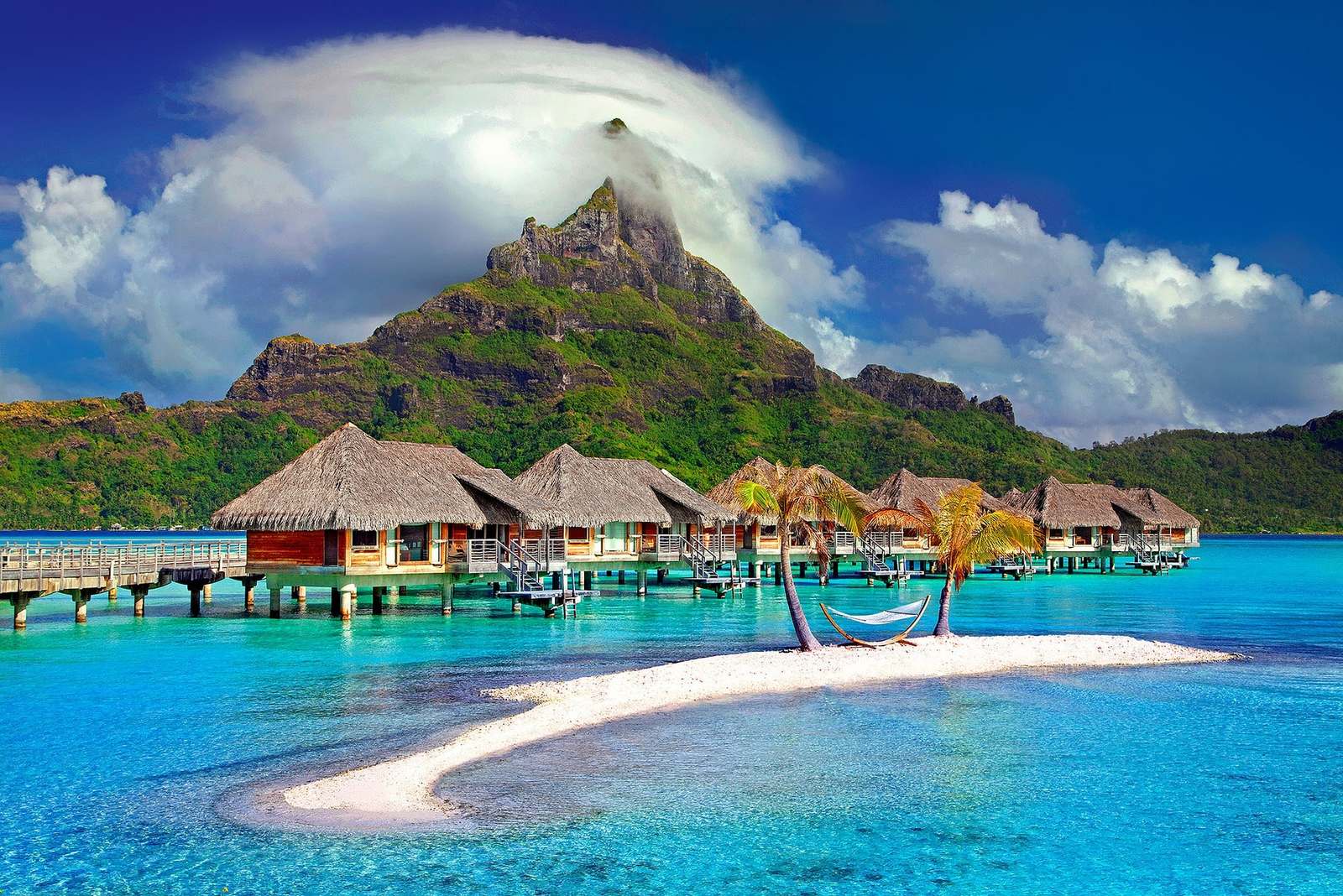 Overwater bungalows and a hammock on a white sandbar below a green volcanic peak